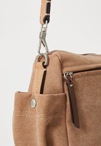 Sac en toile beige avec une finition texturée, doté d'une fermeture zippée, d'accents en cuir, d'une poche latérale et de quincaillerie en métal pour l'attache.