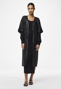 Kimono lung negru din paiete, cu mâneci largi, purtat peste o rochie neagră ajustată. Sandale plate negre. Textură netedă și accente strălucitoare.