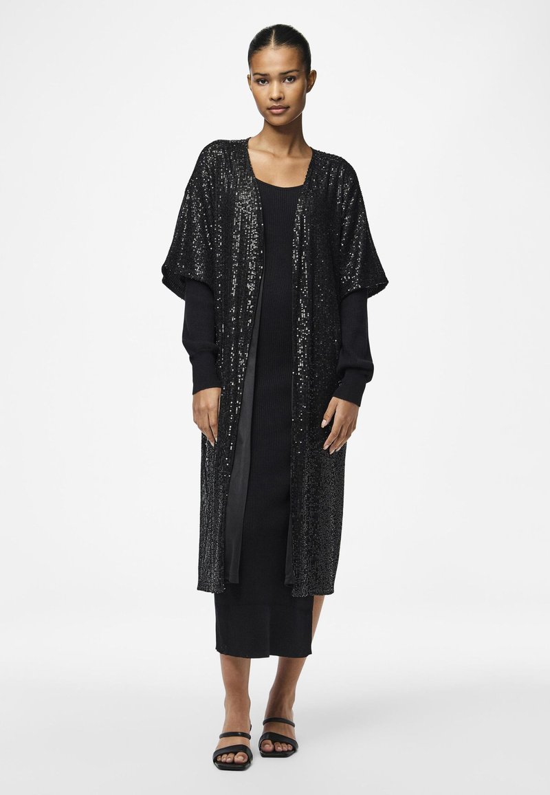 Kimono lung negru din paiete, cu mâneci largi, purtat peste o rochie neagră ajustată. Sandale plate negre. Textură netedă și accente strălucitoare.