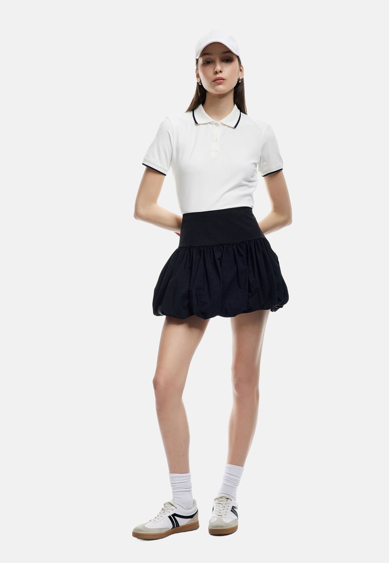 Vit polo skjorta med svarta detaljer, tillsammans med en svart plisserad kjol. Modellen bär en vit keps och sneakers med svarta och beige accenter.