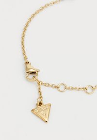 Gouden kettingarmband met ovale schakels en een kreeftslot, versierd met een driehoekige hanger gegraveerd met "GUESS." Gladde textuur.