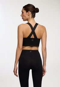 Top sportivo nero con spalline incrociate, realizzato in tessuto elasticizzato, presenta una texture liscia e un logo del marchio sulla parte posteriore.