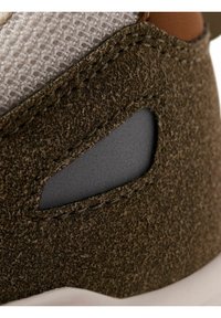 Gros plan sur du daim marron texturé avec un détail découpé noir et un tissu blanc tissé au-dessus, partie d’un modèle de chaussure ou de basket.