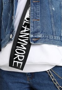 Giacca di jeans con dettagli strappati, indossata sopra un felpa bianca. Cintura nera con bold white text "ANYMORE" in evidenza.