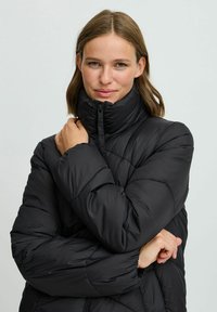 Chaqueta de plumas negra con cuello alto, que presenta un diseño acolchado y una textura suave. El cierre frontal y el ajuste holgado son detalles notables.