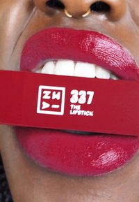 Röd läppstift med glossig finish, visad i munnen, hålls med en rektangulär förpackning märkt "ZW 337 THE LIPSTICK" i vit text.