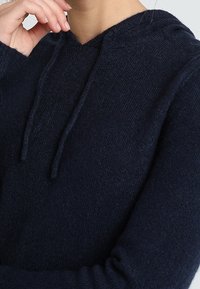 Marinblå hoodiesweater i mjuk stickad tyg, med v-ringning, dragsko och ribbade ärmslut för en snug passform. Nära detalj av texturen.