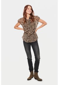 Bluse mit Leopardenmuster und kurzen Ärmeln, mit Rüschendetails an der Vorderseite. Kombination mit dunklen, schmalen Jeans und braunen Stiefeletten. Einfarbiger weißer Hintergrund.