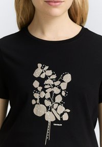 Svart t-shirt med en beige abstrakt växtdesign och ljusa sömdekorationer. Märkets namn "TOM TAILOR" visat längst ner.