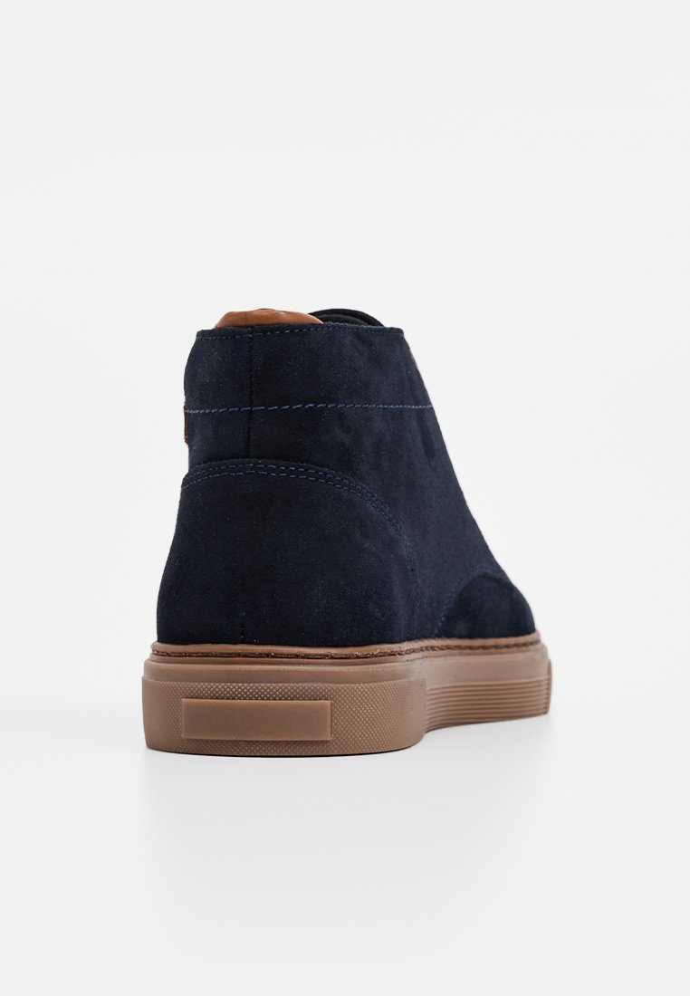 Marineblaues Wildleder-Ankle-Boot mit einem cognacfarbenen Lederakzent an der Ferse, Gummisohle und kontrastierenden blauen Nähten. Glatte Textur und elegantes Design.