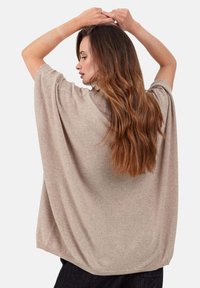 Pull oversize beige en maille avec une texture douce, des manches larges et un ourlet arrondi, porté de profil avec des cheveux flottants.