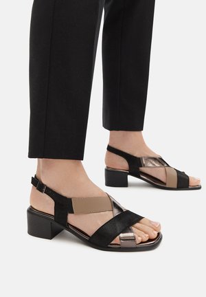 Black and metallic strappy sandals with a low block heel, featuring a mix of materials and colors, showcasing cross straps and ankle support.