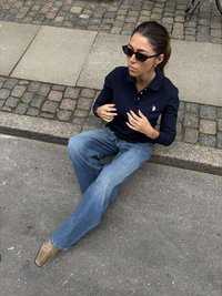 Tricou polo bleumarin, blugi largi bleu deschis și pantofi bej cu vârf ascuțit. Accesorii includ ochelari de soare. Stând pe o suprafață de piatră cubică.