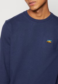 Navy blue sweatshirt with a round neckline, featuring a small embroidered orange car with a turquoise canoe on the left chest. Soft fabric texture.