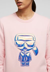 Felpa rosa con un grafico brillante e multicolore di un personaggio con occhiali da sole, tuta dettagliata e coda di cavallo, centrato sul petto.