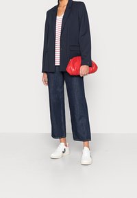 Person iført marineblå blazer, hvid og rød stribet skjorte, mørke brede jeans, hvide sneakers, holder en rød clutch-tasker, stående på hvidt gulv.