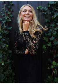 Fekete csipkeruha hosszú ujjakkal, virágmintákkal díszítve, sötét fa háttér előtt, zöld borostyán motívumokkal.