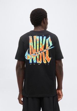 Homme portant un t-shirt noir montrant le logo coloré "Just Do It" de Nike dans le dos sur un fond clair uni.