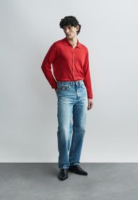 Red button-up shirt, light blue denim jeans, black shoes. Simple design with standard collar and wooden buttons, slightly faded denim texture.