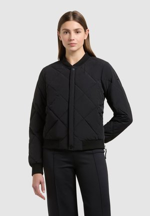 Nő fekete steppelt bomber dzsekiben, bordázott mandzsettával és gallérral, egy egyszerű világos háttér előtt áll.