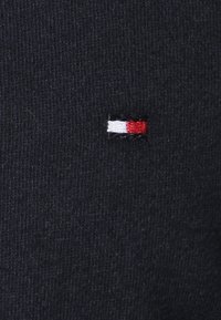 Pequeño logotipo rectangular bordado en rojo, blanco y negro sobre un fondo de tela acanalada de color azul marino oscuro.