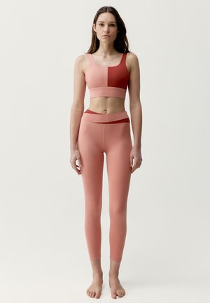 Femme debout pieds nus portant un brassière de sport bicolore rose et rouge et des leggings taille haute assortis sur un fond uni.