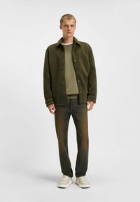 Olivgrüner Fleece-Jacke mit Knopfleiste, über einem hellgrünen Sweatshirt getragen. Kombiniert mit dunkelblauen Jeans und neutralen Sneakers.