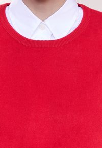 Suéter de punto rojo con un escote redondo y puños de canalé, superpuesto a una camisa blanca con cuello. Textura suave y costura uniforme en toda la prenda.