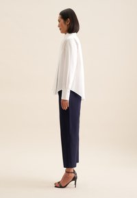 Blusa bianca a maniche lunghe con orlo alto-basso; abbinata a pantaloni tailleur blu navy; la modella indossa tacchi a strappi neri; sfondo chiaro.