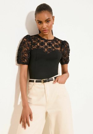 Femme aux cheveux foncés portant un haut noir avec des manches bouffantes en dentelle florale transparente et un pantalon crème taille haute avec une ceinture noire.