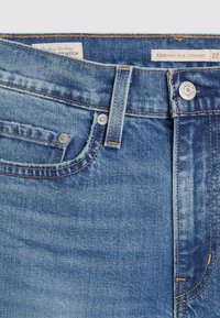 Gros plan sur un jean denim bleu taille haute, coupe droite, avec passants de ceinture visibles, poche avant, rivets métalliques et fermeture par bouton.