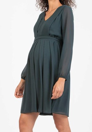 Gravid kvinde iført en mørkegrøn langærmet knælængde kjole med V-hals og plisseret nederdel.