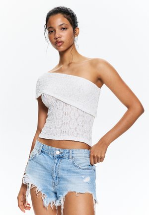 Witte strapless top met gestructureerde stof en geplooide details, gecombineerd met lichtblauwe distressed denimshorts met franjes.
