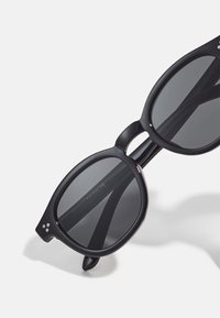 Schwarze Sonnenbrille mit runden Rahmen. Dunkle Gläser und dicke, glatte Bügel. Auffällige Details sind kleine silberne Punkte am Rahmen.