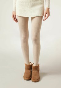 Des collants beiges à motifs avec un design texturé, associés à des bottines en daim marron doublées de peluche. Le modèle porte une jupe blanche.