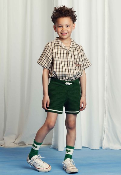 Child wears a brown plaid short-sleeve button-up shirt, green textured shorts with white trim, and green and white patterned socks with sneakers.