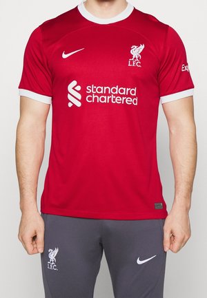 Mann trägt ein rotes Liverpool FC Fußballtrikot mit weißen Logos und dunkelgrauen Nike-Hosen, steht vor einem einfarbigen Hintergrund.