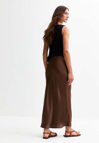 Jupe maxi en satin marron avec une coupe fluide, associée à un haut noir sans manches ajusté et des sandales plates, vue de l'arrière.