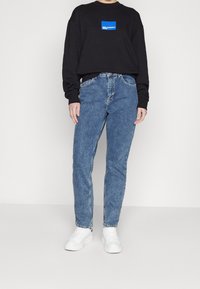 Sudadera negra con logotipo azul, jeans de mezclilla azul y zapatillas blancas. Corte relajado, mangas largas y diseño de cintura alta.