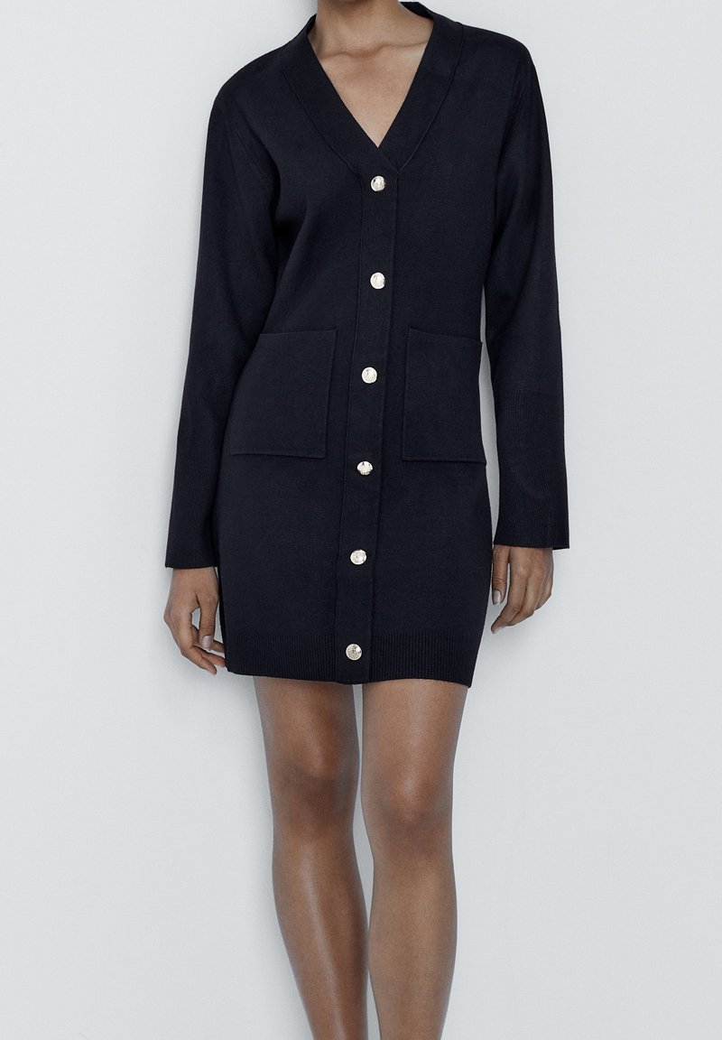 Femme portant un long cardigan noir boutonné avec deux poches avant, col en V et manches longues, debout devant un fond uni.