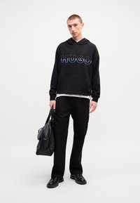 Svart hettegenser med "HUGO" logo; kengurulomme. Svart bukse, glansete svarte sko og en svart skinnveske. Enkelt, uformelt design.