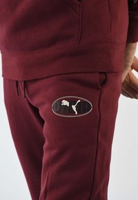 Pantalones deportivos granate hechos de tela suave, con un parche de logo redondeado con acentos en blanco y negro. Cintura elástica, con bolsillos laterales visibles.