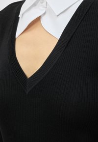 Κοντινό πλάνο ενός μαύρου ριμπ πλεκτού πουλόβερ με V-λαιμόκοψη που φοριέται πάνω από ένα λευκό πουκάμισο με γιακά, αφήνοντας ακάλυπτο μέρος του δέρματος στην περιοχή του στήθους.