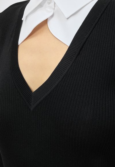 Gros plan d'un pull noir côtelé à col en V porté par-dessus une chemise blanche à col, révélant un peu de peau au niveau de la poitrine.