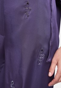 Purple satin fabric featuring beaded snake designs in silver and black, located on the side near a hand. The texture is smooth and glossy.