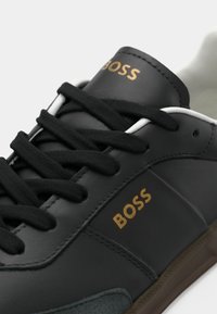 Baskets en cuir noir avec lacets noirs, marquage doré "BOSS" sur la languette et le côté, bordure blanche et semelle marron.
