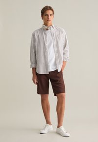 Camicia a righe in marrone chiaro e bianco, abbinata a pantaloni corti marroni e sneakers bianche. Presenta una tasca e maniche arrotolate.