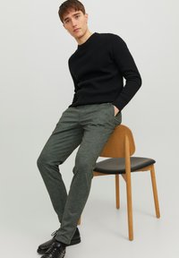 Maglione nero a coste, pantaloni verdi texture e scarpe nere. Modello seduto su una sedia di legno con sedile nero. Sfondo chiaro e uniforme.
