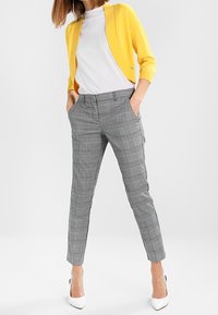 Geel vest over een wit T-shirt, gecombineerd met grijze geruite broek en witte hakken. Het vest is lichtgewicht en de broek is op maat gemaakt.