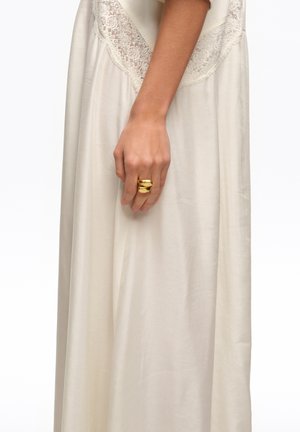 Mano apoyada sobre un vestido color crema con detalles de encaje, llevando un llamativo anillo dorado grueso en el dedo medio.